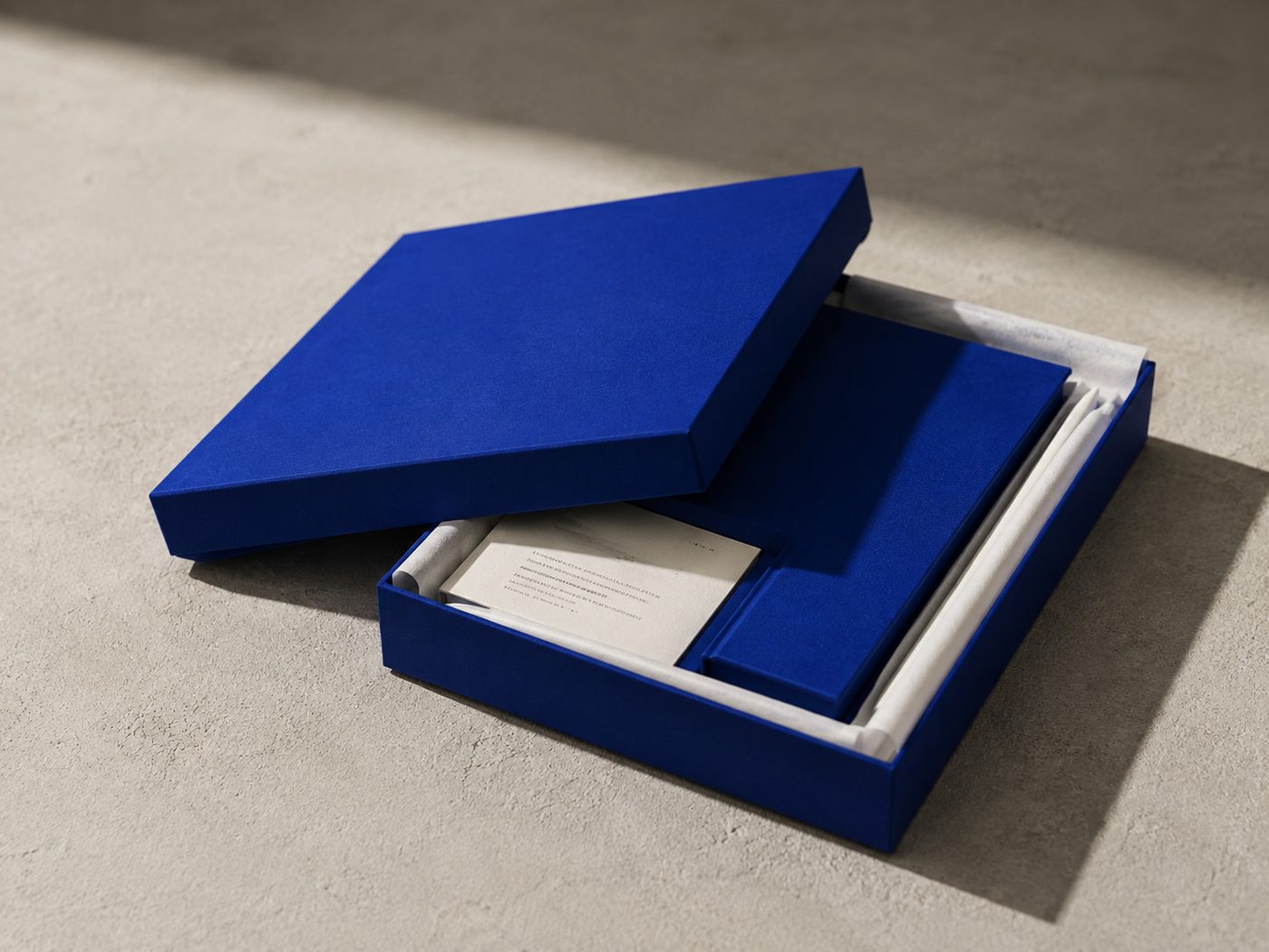The presentation box in matte blue, lid ajar, book and pen inside on off-white tissue.